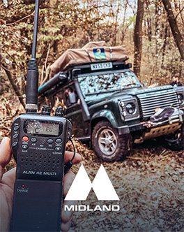 Talkie-walkie Midland Alan tenu en main devant un 4x4 équipé d'une tente de toit en forêt